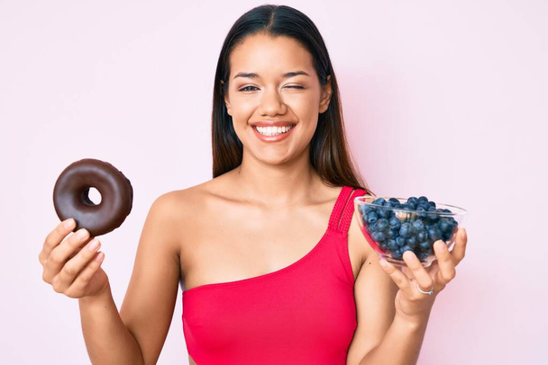 Young beautiful latin girl wearing swimsuit holding donut and blueberries winking looking at the camera with sexy expression, cheerful and happy face.  - Foto, Imagem