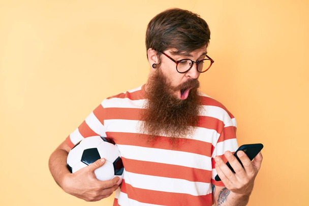 Przystojny młody rudy mężczyzna z długą brodą trzymający piłkę nożną patrząc na smartfona przerażony i zszokowany zaskakującą i zdumioną ekspresją, strachem i podnieconą twarzą.  - Zdjęcie, obraz