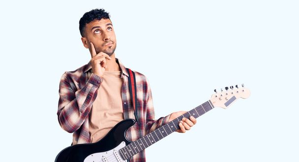 Νεαρός Ισπανός που παίζει ηλεκτρική κιθάρα με σοβαρό πρόσωπο σκεπτόμενος την ερώτηση με το χέρι στο πηγούνι, σκεπτόμενος την μπερδεμένη ιδέα  - Φωτογραφία, εικόνα