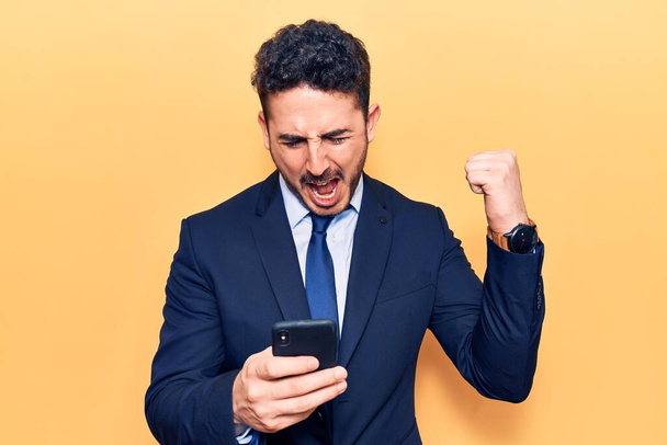 Young hispanic man wearing suit using smartphone screaming proud, celebrating victory and success very excited with raised arms  - Photo, Image