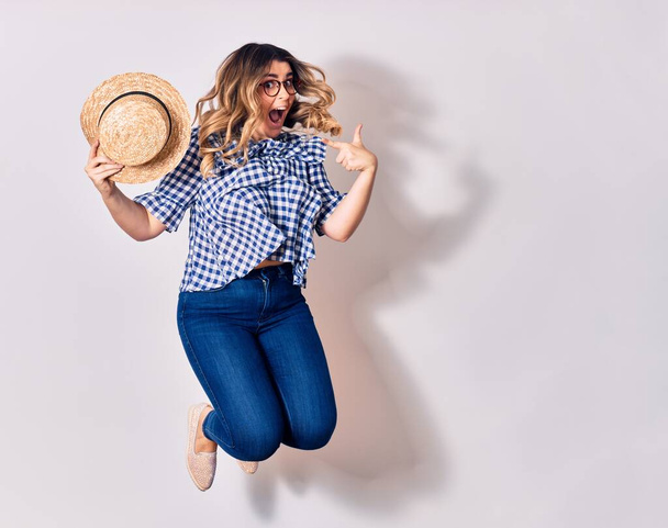 Young beautiful blonde woman wearing glasses holding hat smiling happy. Jumping with smile on face over isolated white background. - Фото, зображення