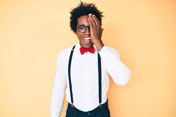 Handsome african american man with afro hair wearing hipster elegant look covering one eye with hand, confident smile on face and surprise emotion.  - Fotoğraf, Görsel