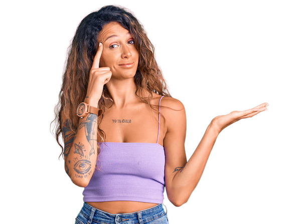Jeune femme hispanique avec tatouage portant des vêtements décontractés confus et agacés avec la paume ouverte montrant l'espace de copie et pointant du doigt vers le front. Penses-y..  - Photo, image