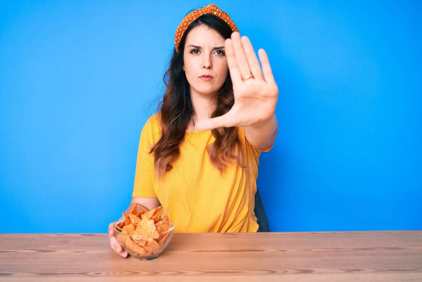 Joven hermosa morena sentada en la mesa comiendo nachos patatas fritas con la mano abierta haciendo stop sign con expresión seria y segura, gesto de defensa  - Foto, Imagen