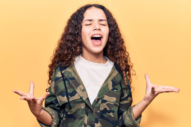 Beautiful kid girl with curly hair wearing camouflage jacket crazy and mad shouting and yelling with aggressive expression and arms raised. frustration concept.  - Фото, изображение