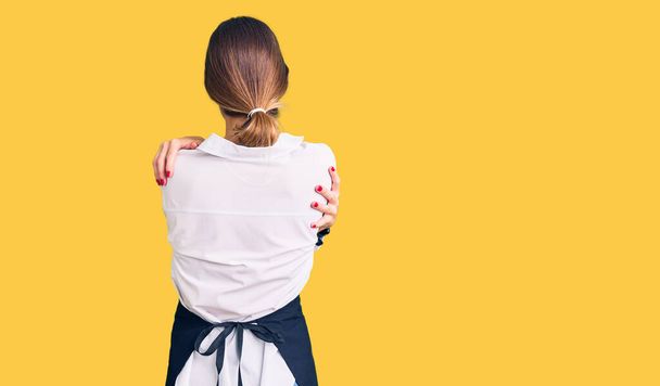 Beautiful brunette young woman wearing professional waitress apron hugging oneself happy and positive from backwards. self love and self care  - Foto, imagen