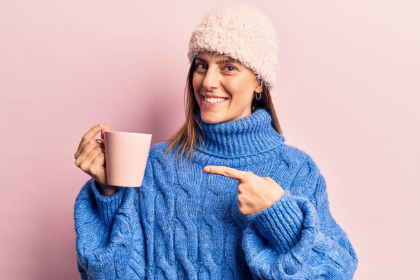 Молодая красивая женщина в зимней одежде держа кофе улыбаясь счастливо указывая рукой и пальцем  - Фото, изображение