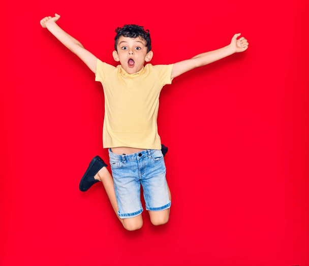 Αξιολάτρευτο παιδί που φοράει καθημερινά ρούχα πηδώντας πάνω από απομονωμένο κόκκινο φόντο - Φωτογραφία, εικόνα