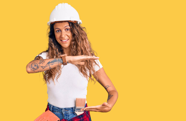Jeune femme hispanique avec tatouage portant un casque et des vêtements de constructeur gesticulant avec des mains montrant grand et grand signe de taille, symbole de mesure. souriant en regardant la caméra. concept de mesure.  - Photo, image
