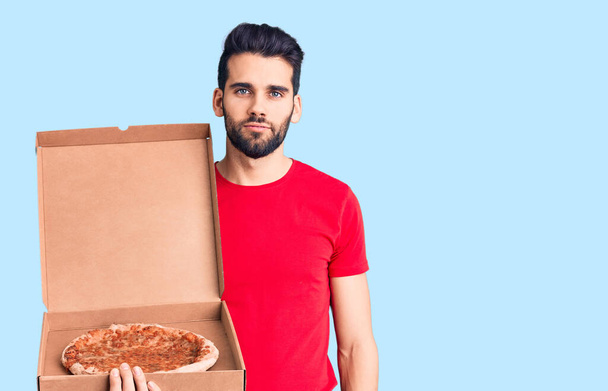 Junger gutaussehender Mann mit Bart hält Lieferkarton mit italienischem Pizza-Denken und nüchternem Gesichtsausdruck und wirkt selbstbewusst  - Foto, Bild