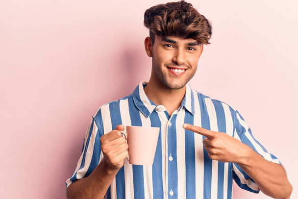 Young hispanic man holding coffee smiling happy pointing with hand and finger  - Fotografie, Obrázek