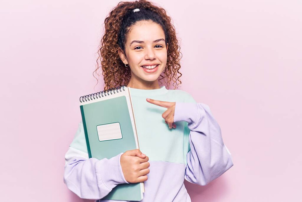 Όμορφο κοριτσάκι με σγουρά μαλλιά κρατώντας σημειωματάριο χαμογελώντας χαρούμενος δείχνοντας με το χέρι και το δάχτυλο  - Φωτογραφία, εικόνα