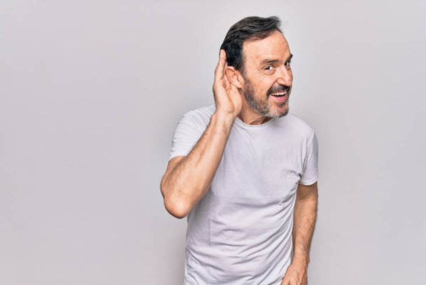 Homem bonito de meia idade vestindo camiseta casual em pé sobre fundo branco isolado sorrindo com a mão sobre a orelha ouvindo e ouvindo rumores ou fofocas. Conceito de surdez
. - Foto, Imagem