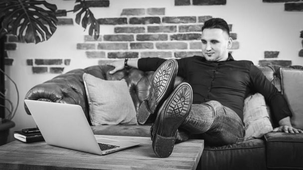 Multitasking. A handsome guy in a black shirt and jeans is lying on the couch, next to him on the table turned on the laptop, he works in the office. Remote work. Black and white photo - Foto, imagen