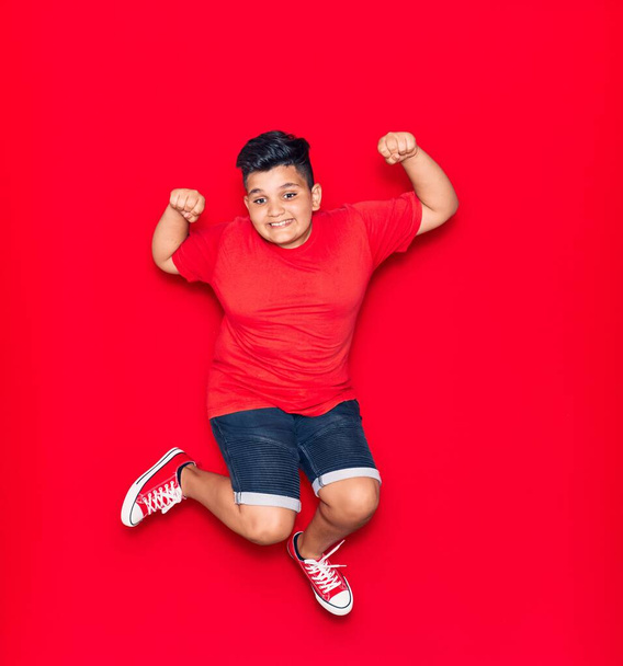 Adorable kid wearing casual clothes smiling happy. Jumping with smile on face doing strong gesture with arms over isolated red background - Φωτογραφία, εικόνα