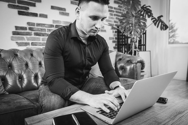 Multitasking. Ein gutaussehender Typ in schwarzem Hemd und Jeans liegt auf der Couch, neben ihm auf dem Tisch ein Laptop, er arbeitet im Büro. Fernbedienung. Schwarz-Weiß-Foto - Foto, Bild