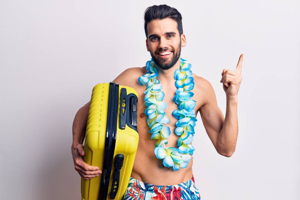 Young handsome man with beard wearing swimwear and hawaiian lei holding plane cabin bag smiling happy pointing with hand and finger to the side  - Foto, immagini