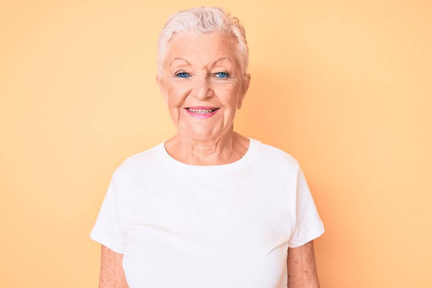 Belle femme âgée aux yeux bleus et aux cheveux gris portant un t-shirt blanc classique sur fond jaune avec un sourire heureux et cool sur le visage. chanceux.  - Photo, image