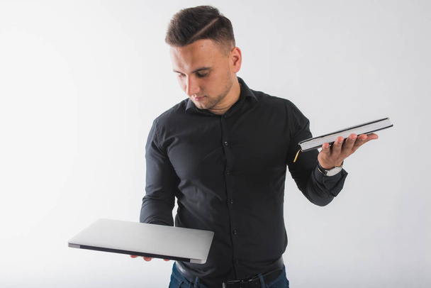 Ένας νεαρός άνδρας με ένα μαύρο πουκάμισο, κρατώντας ένα φορητό υπολογιστή και ένα βιβλίο και να αποφασίσει τι να επιλέξει, απομονώνονται σε λευκό φόντο, υπάρχει μια θέση για κείμενο - Φωτογραφία, εικόνα