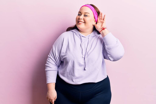 Young plus size woman wearing sportswear smiling with hand over ear listening and hearing to rumor or gossip. deafness concept.  - Foto, afbeelding