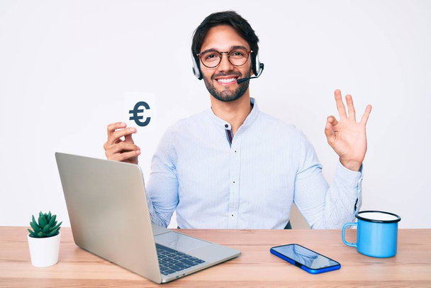 Knappe Spaanse man die op kantoor werkt met een eurospandoek en met z 'n vingers goed gebarentaal maakt, glimlachend vriendelijk gebaar en uitstekend symbool  - Foto, afbeelding