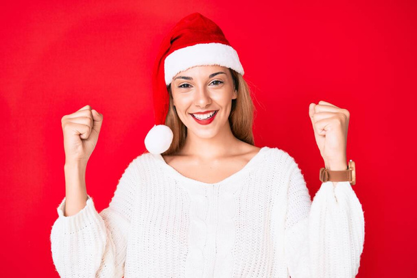 Młoda brunetka w świątecznym kapeluszu krzyczy dumny, świętując zwycięstwo i sukces bardzo podekscytowany z podniesionymi ramionami  - Zdjęcie, obraz