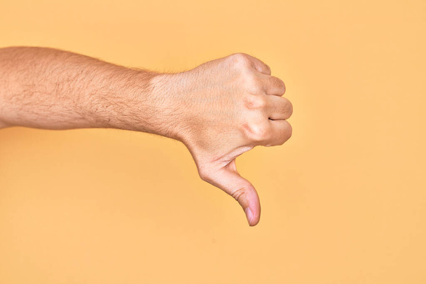 Ruka bělocha, mladý muž ukazující prsty na izolovaném žlutém pozadí, ukazující palce dolů odmítavé gesto, nesouhlas a negativní znamení - Fotografie, Obrázek