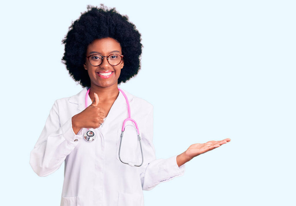 Mladá africká Američanka v lékařském kabátě a stetoskopu ukazující dlaň a dělá v pořádku gesto s palci nahoru, usmívá se šťastný a veselý  - Fotografie, Obrázek