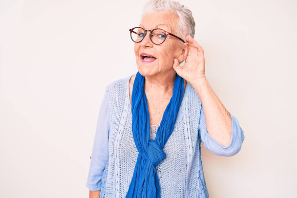 青い目と灰色の髪をしたシニア美しい女性は、カジュアルなセーターとスカーフを着て、耳を傾け、噂やゴシップに耳を傾けながら手で笑っています。聴覚障害の概念.  - 写真・画像