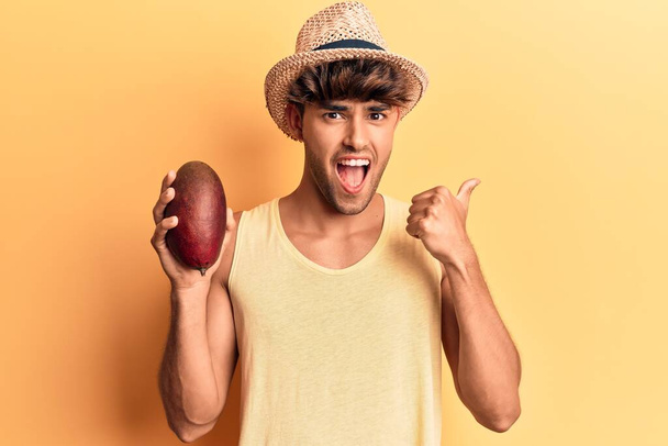 Joven hispano con sombrero sosteniendo mango apuntando con el pulgar hacia el costado sonriendo feliz con la boca abierta  - Foto, Imagen