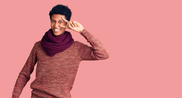 Giovane uomo afroamericano indossa casual maglione invernale e sciarpa facendo simbolo di pace con le dita sul viso, sorridente allegro mostrando la vittoria  - Foto, immagini