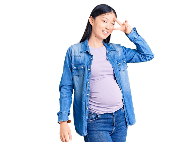 Молодая красивая китаянка беременная ждет ребенка делая символ мира с пальцами на лице, улыбаясь весело показывая победу  - Фото, изображение