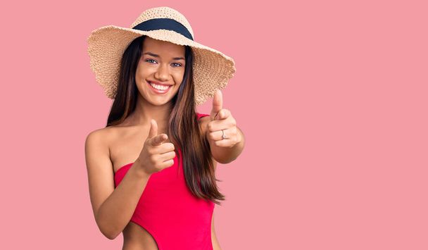 Молодая красивая латинская девушка в купальниках и летней шляпе, указывающая пальцами на камеру со счастливым и смешным лицом. хорошая энергия и вибрации.  - Фото, изображение
