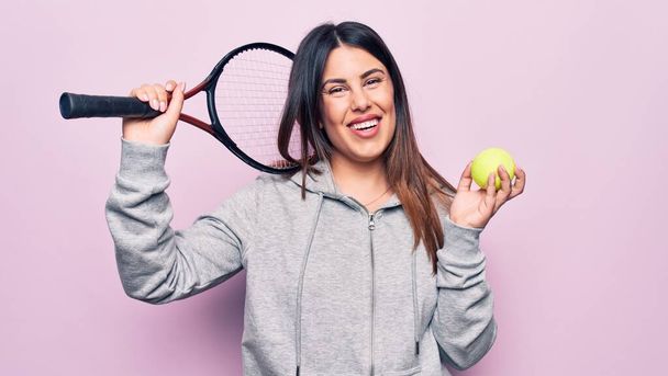 Joven deportista hermosa jugando al tenis usando raqueta y pelota sobre fondo rosa mirando positiva y feliz de pie y sonriendo con una sonrisa confiada mostrando los dientes - Foto, Imagen