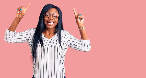 若いアフリカ系アメリカ人の女性は、カジュアルな服や眼鏡を身に着けている笑顔と驚きと指で指摘し、腕を上げた.  - 写真・画像