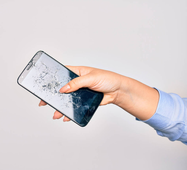 Hand of caucasian young woman holding broken smartphone showing craked screen over isolated white background - Photo, Image