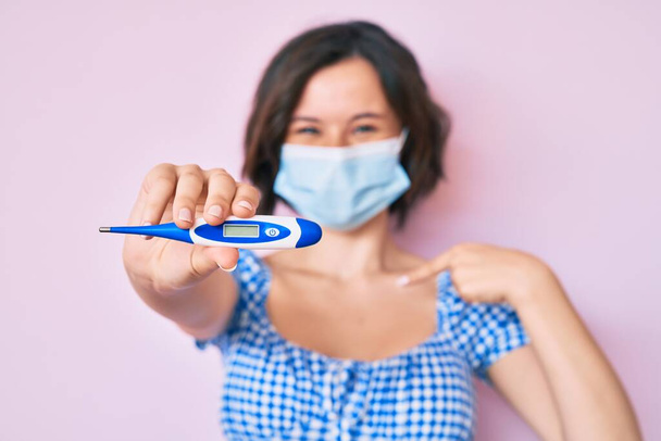Young beautiful woman wearing medical mask holding holding thermometer pointing finger to one self smiling happy and proud  - Photo, Image