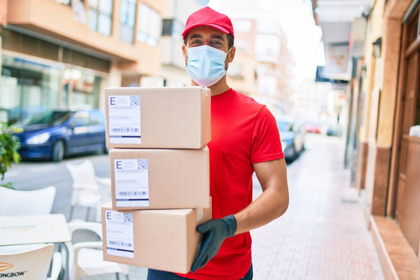 Jonge bezorger in uniform en coronavirus bescherming medisch masker met kartonnen pakjes op straat. - Foto, afbeelding