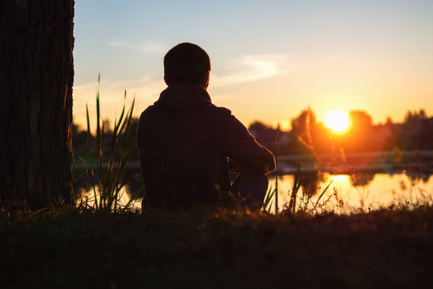 Junger einsamer Mann, der bei Sonnenuntergang am See sitzt. Genießen Sie die Aussicht - Foto, Bild