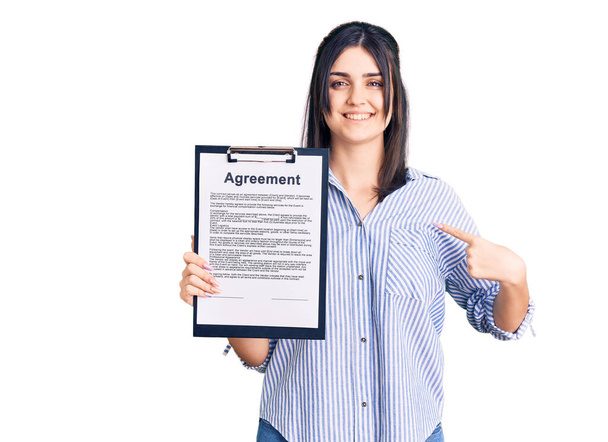 Jeune belle fille tenant presse-papiers avec document d'accord pointant du doigt un sourire heureux et fier  - Photo, image