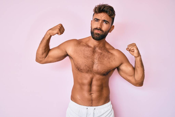 Joven hombre hispano de pie sin camisa mostrando los músculos de los brazos sonriendo orgulloso. concepto de fitness.  - Foto, imagen