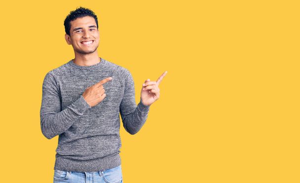 Der hispanische gutaussehende junge Mann trägt einen lässigen Pullover und lächelt in die Kamera, die mit zwei Händen und Fingern zur Seite zeigt..  - Foto, Bild