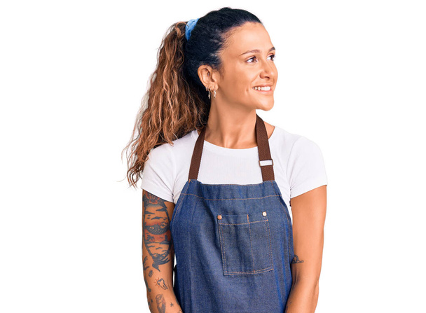 Mujer hispana joven con tatuaje usando delantal de peluquero y guantes mirando hacia otro lado con sonrisa en la cara, expresión natural. riendo confiado.  - Foto, Imagen