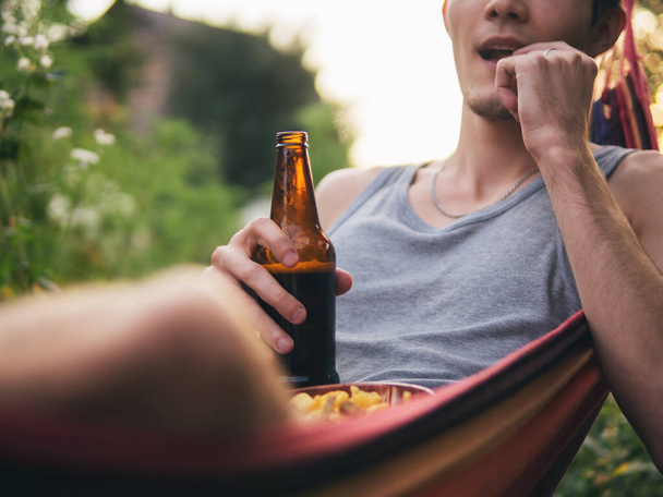 Joven descansando al aire libre en una hamaca y bebiendo cerveza con galletas y patatas fritas - Foto, Imagen