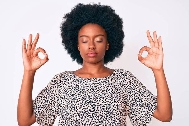 Mladá Afroameričanka v neformálním oblečení odpočívá a usmívá se se zavřenýma očima a prsty provádí meditační gesto. koncepce jógy.  - Fotografie, Obrázek