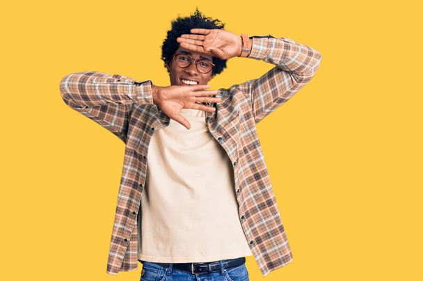 Knappe Afro-Amerikaanse man met afro-haar in casual kleding en een bril die vrolijk glimlacht en gluurt naar een boe met wijzers die hun gezicht laten zien. verrast en verlaten  - Foto, afbeelding