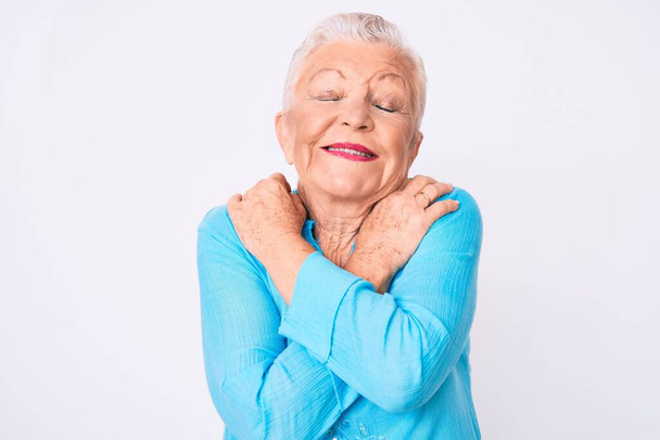 Senioren mooie vrouw met blauwe ogen en grijs haar dragen zomerkleren knuffelen zichzelf gelukkig en positief, glimlachend zelfverzekerd. zelfliefde en zelfzorg  - Foto, afbeelding