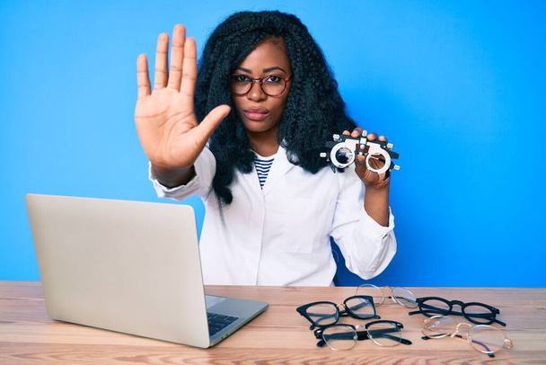 Belle femme africaine tenant des lunettes d'optométrie avec la main ouverte faisant stop signe avec expression sérieuse et confiante, geste de défense  - Photo, image