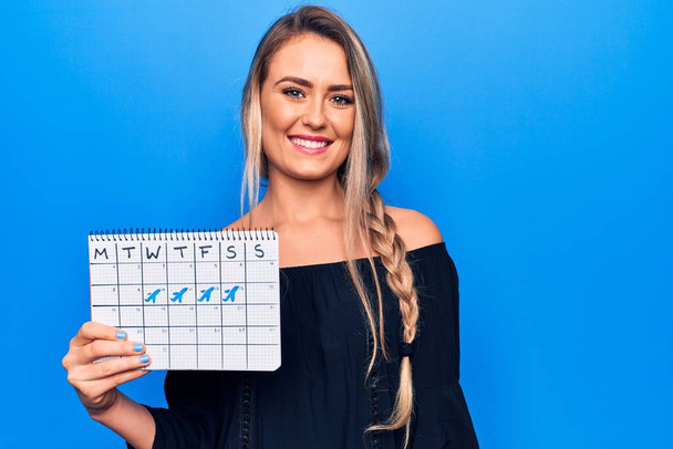 Young beautiful blonde tourist woman holding travel calendar showing vacation week looking positive and happy standing and smiling with a confident smile showing teeth - Photo, Image