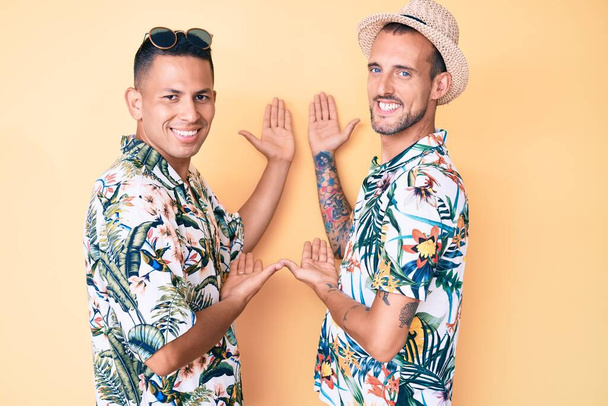 Νέοι γκέι ζευγάρι δύο ανδρών που φορούν καλοκαιρινό καπέλο και χαβανέζικο πουκάμισο προσκαλώντας τους να εισέλθουν χαμογελώντας φυσικά με ανοιχτό χέρι  - Φωτογραφία, εικόνα